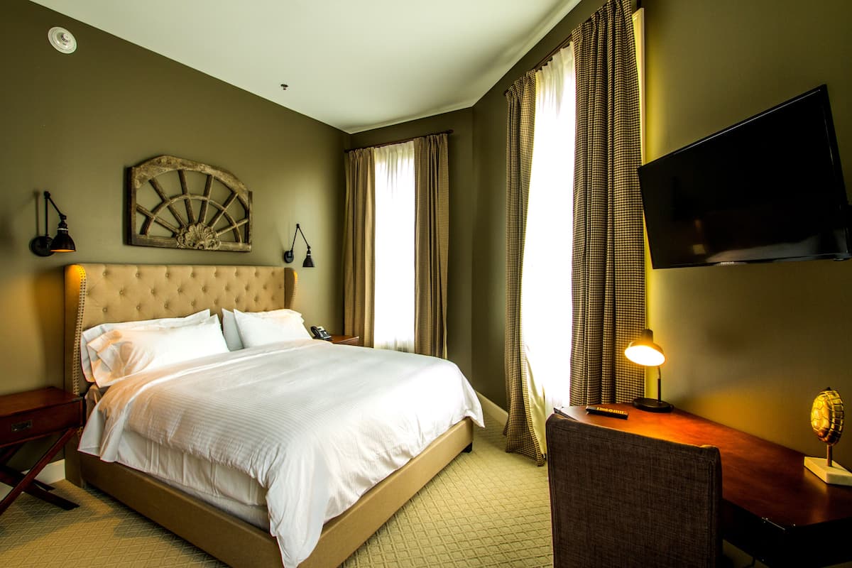 A hotel room with a large bed, a desk with a lamp, a television on the wall, and a decorative wall piece.