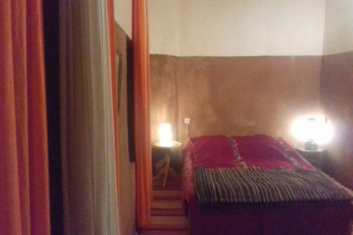 A narrow hallway leading to a bed with a red blanket and striped pillow. There are two bedside lamps and a wall-mounted light.