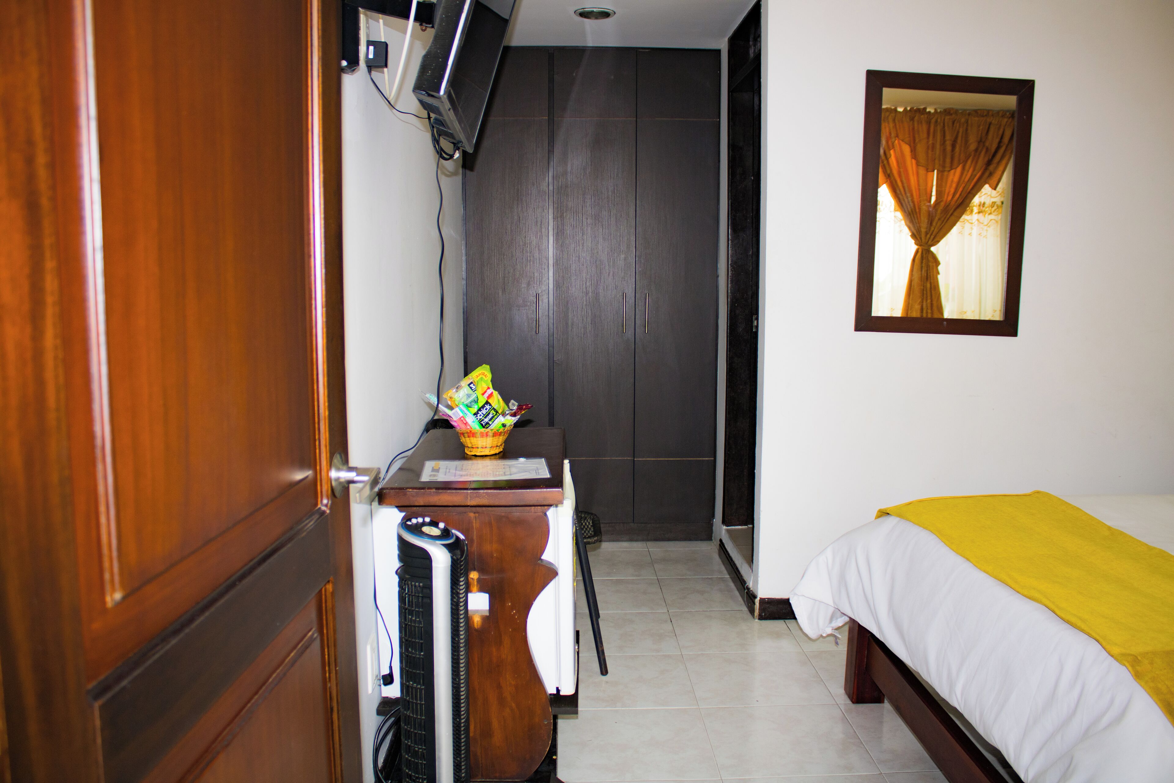 Habitación de hotel con un armario de madera, un escritorio con un cesto de artículos de aseo, una cama con una manta amarilla y una ventana con cortinas.