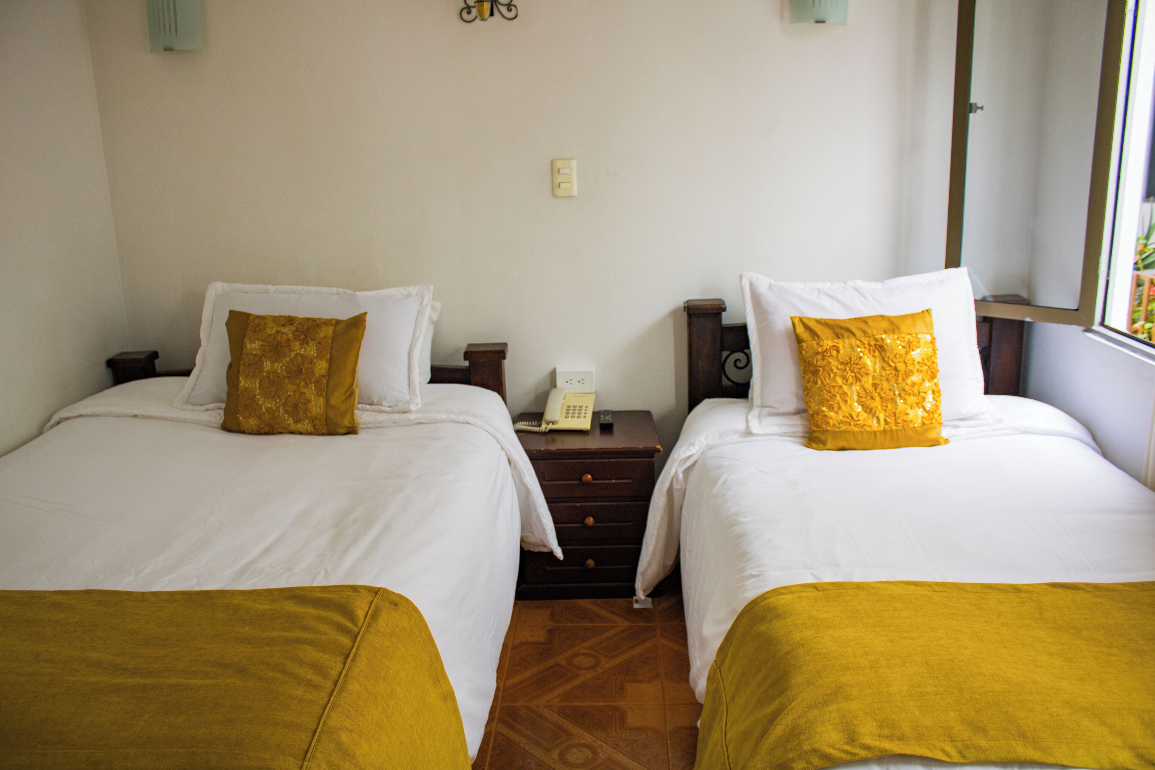 Habitación de hotel con dos camas, cada una con cubrecama blanco y almohada decorativa dorada. Entre las camas, una mesita de noche de madera con un teléfono y una pequeña mesa.