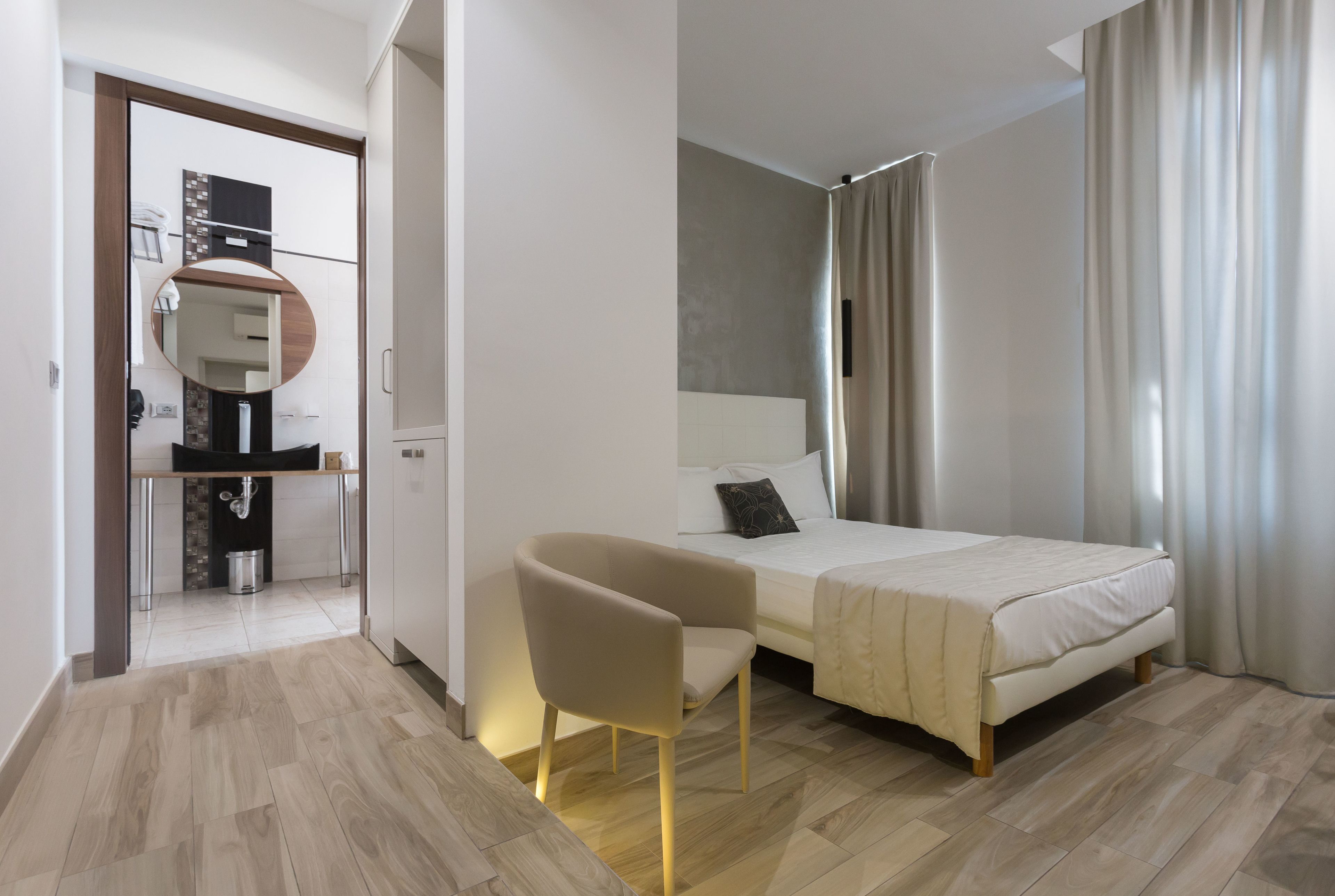 Quarto de hotel moderno com cama, uma cadeira, uma escrivaninha e um banheiro visível por uma porta aberta.