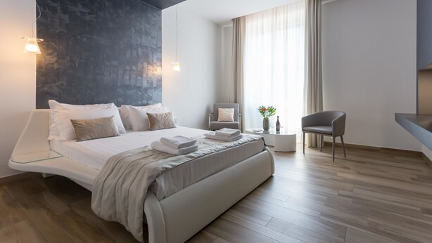 Une chambre à coucher moderne avec un lit aux lignes courbes, une chaise, une petite table et une grande fenêtre avec des rideaux.