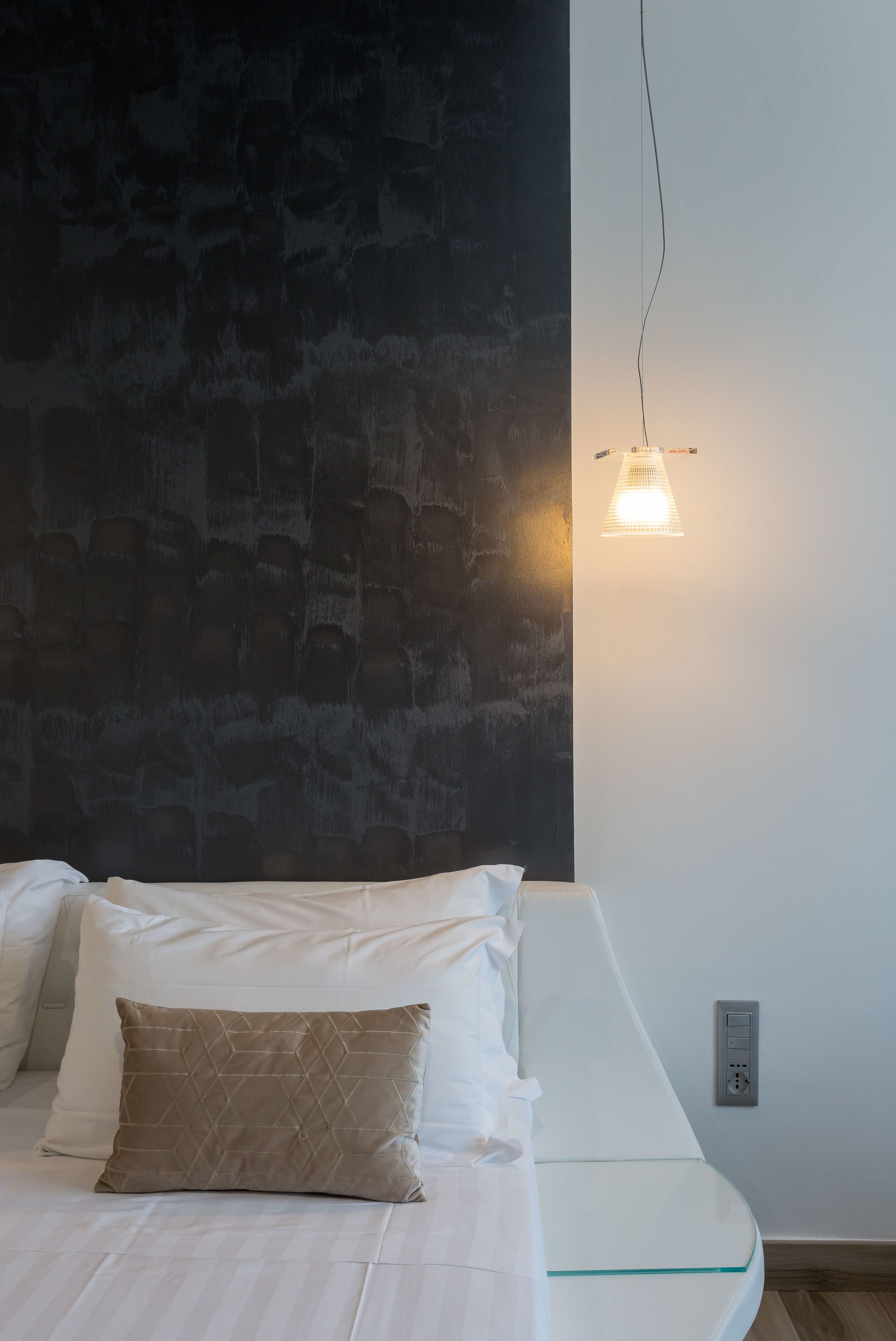 A neatly made bed with white and beige pillows, a glass bedside table, and a wall-mounted light fixture.