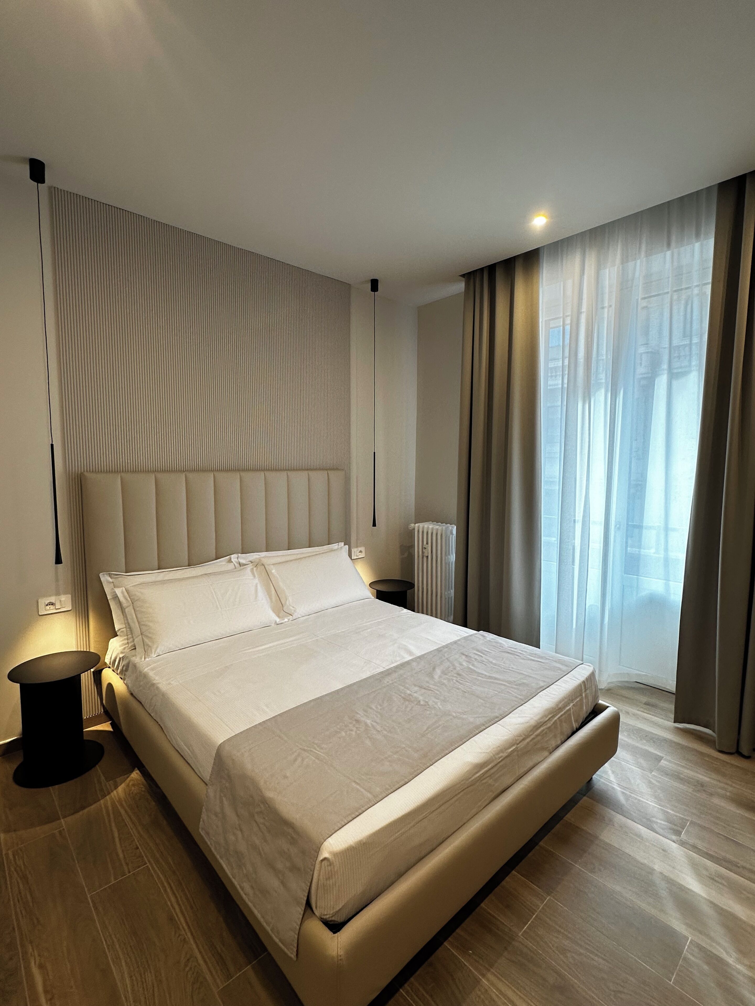 A modern hotel room with a large bed, a bedside table, a radiator, and a window with curtains.