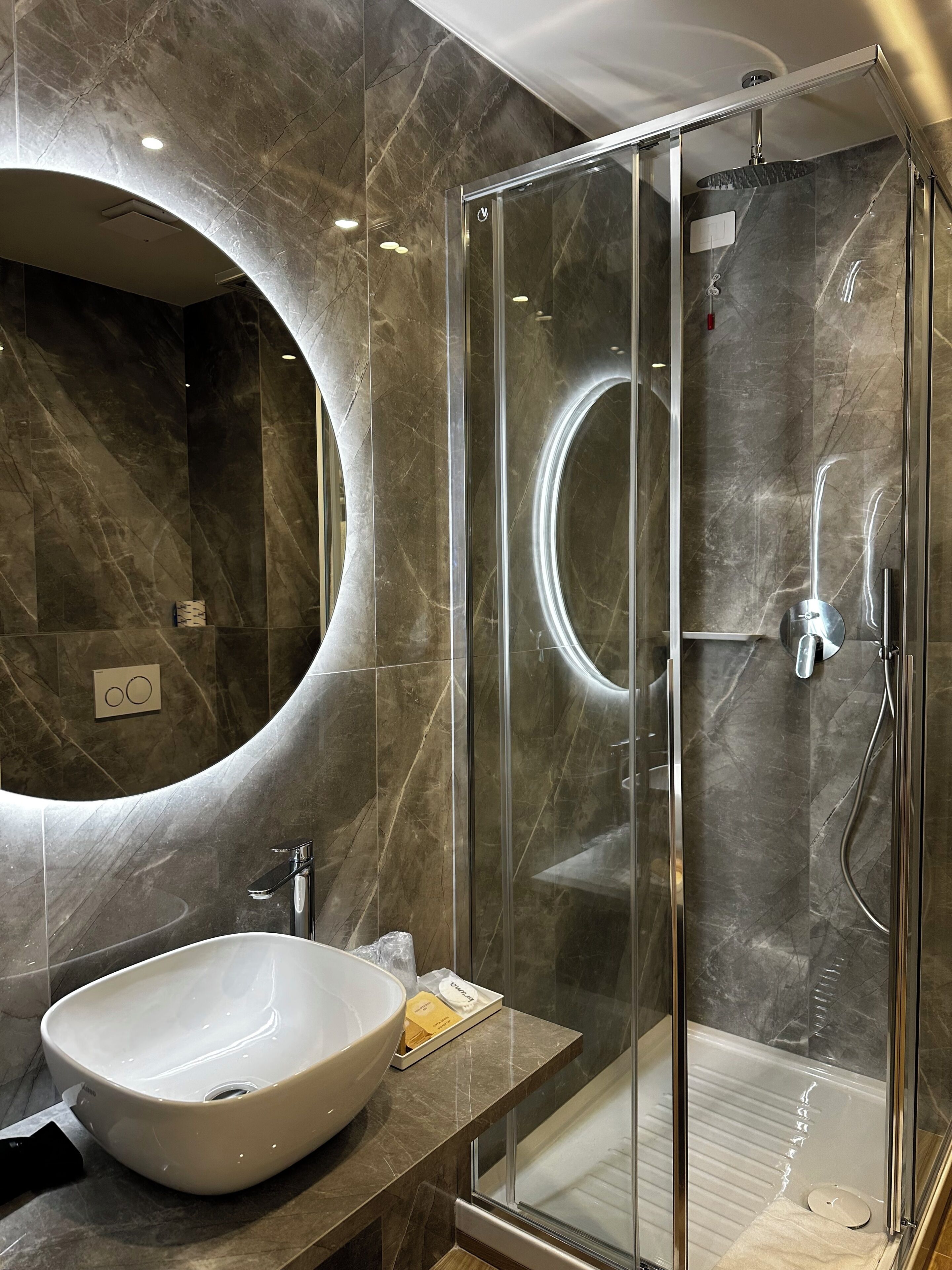 Ein modernes Badezimmer mit einem großen, ovalen Waschbecken, einer gläsernen Dusche und einer Marmorwand mit einem runden Spiegel.