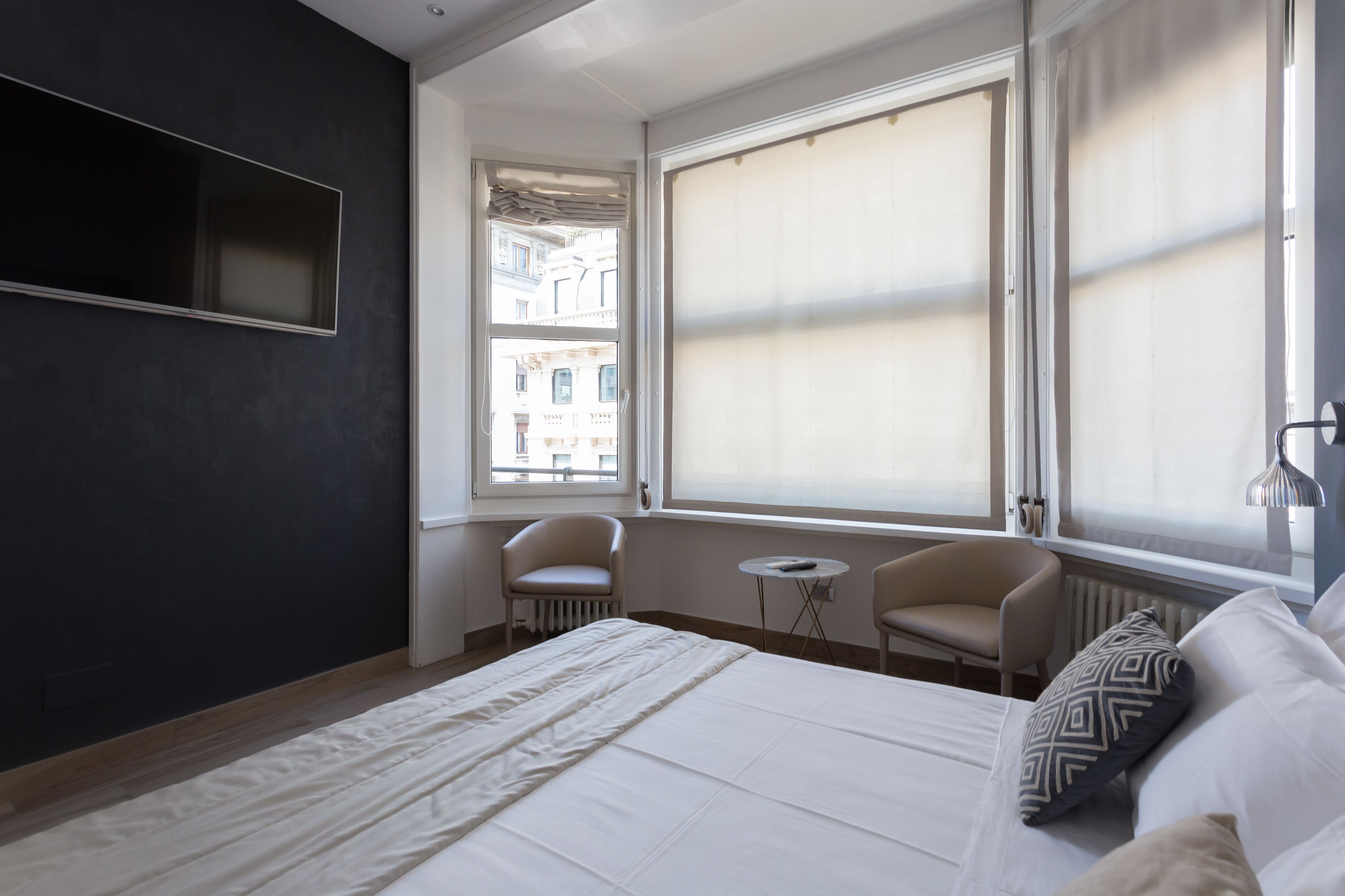 A modern hotel room with a large bed, a small table, two chairs, and a large window with blinds.