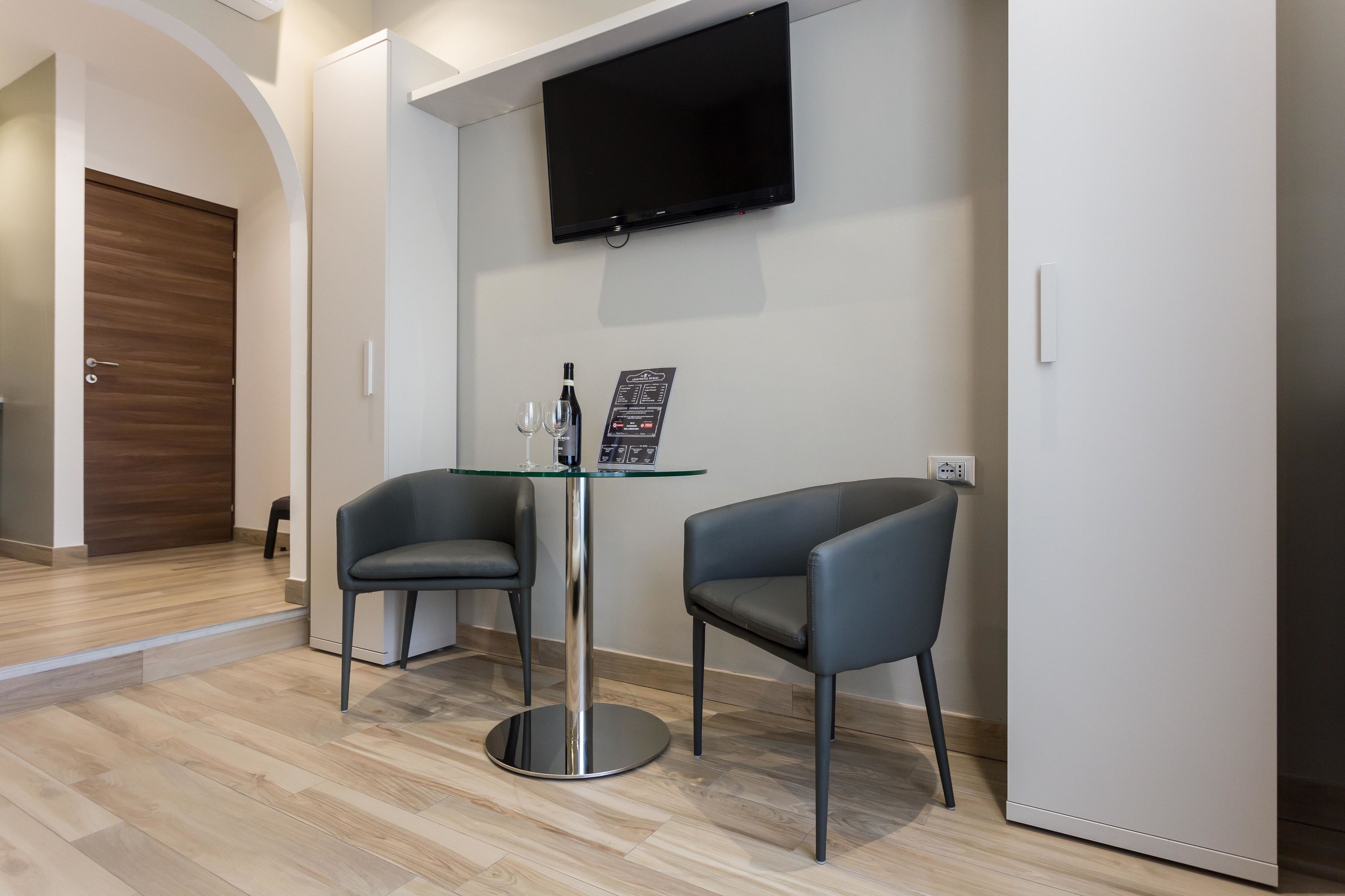 Una habitación moderna con una mesa de vidrio, dos sillas, un televisor de pantalla plana montado en la pared y una puerta de madera.