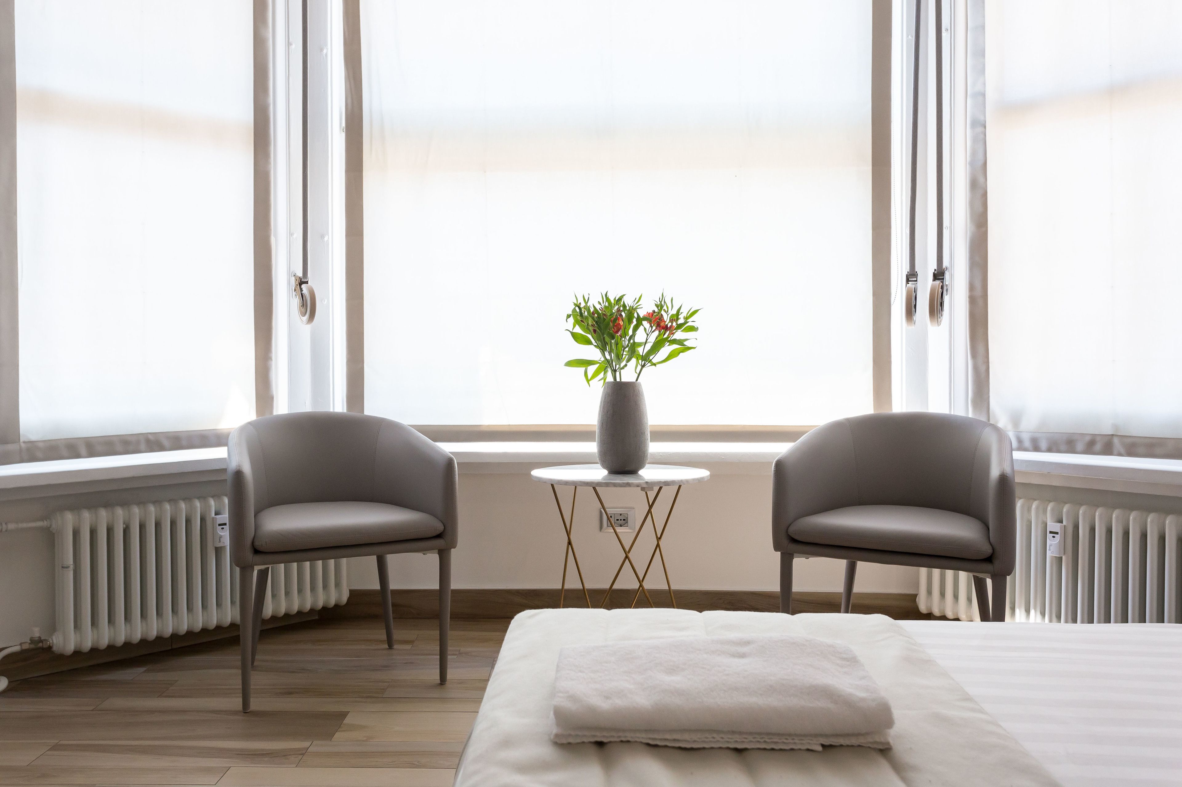 A hotel room with a bed, two chairs, a small table with a vase of flowers, and large windows with curtains.