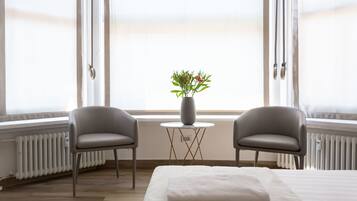 A hotel room with a bed, two chairs, a small table with a vase of flowers, and large windows with curtains.