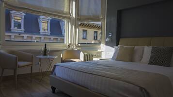 Ein Hotelzimmer mit einem großen Bett, einem kleinen Tisch mit einer Flasche, einem Sessel und Blick auf Gebäude durch große Fenster.