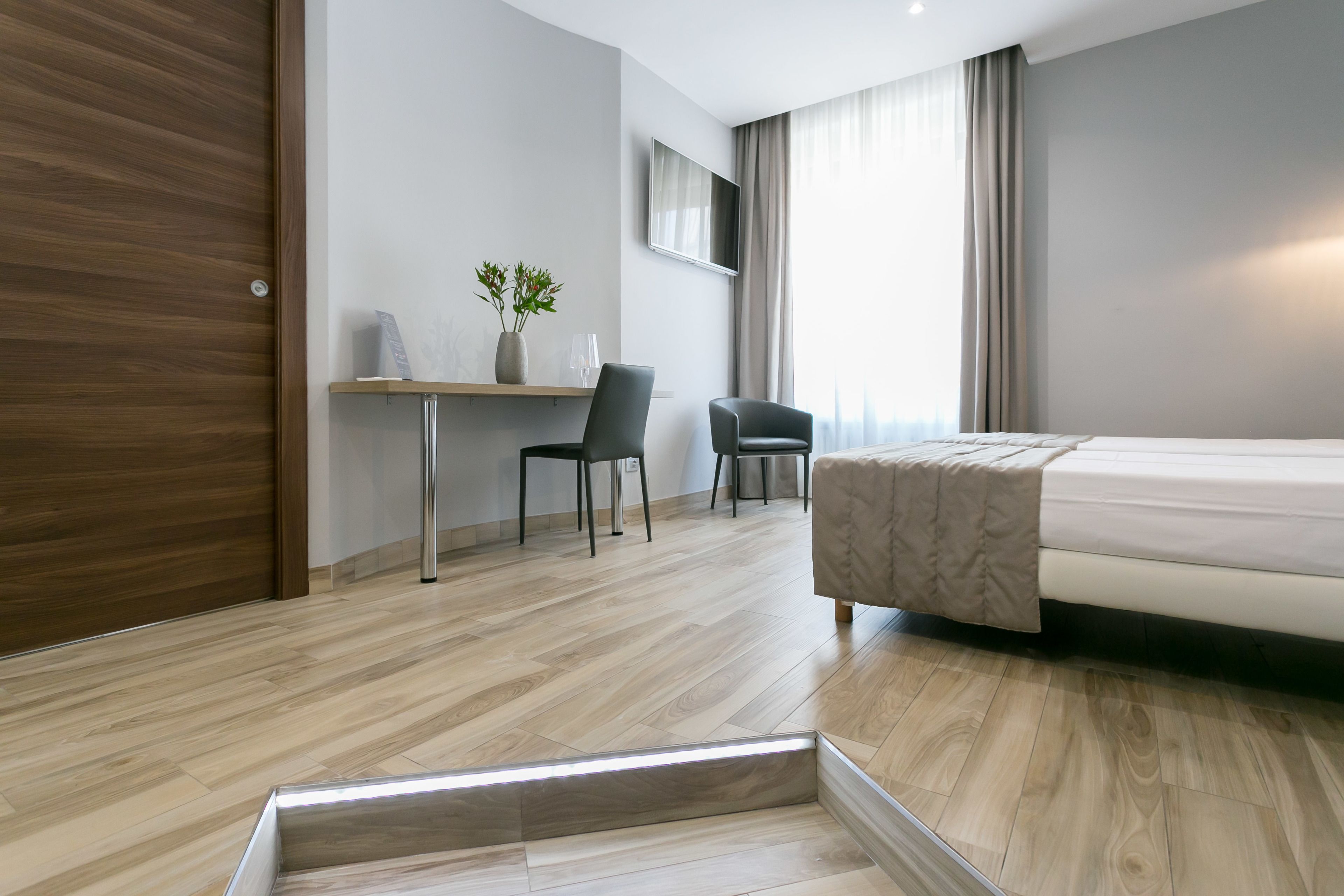 Ein modernes Hotelzimmer mit Holzboden, einem Bett, einem Schreibtisch mit zwei Stühlen, einem Fernseher und einem Fenster mit Vorhängen.