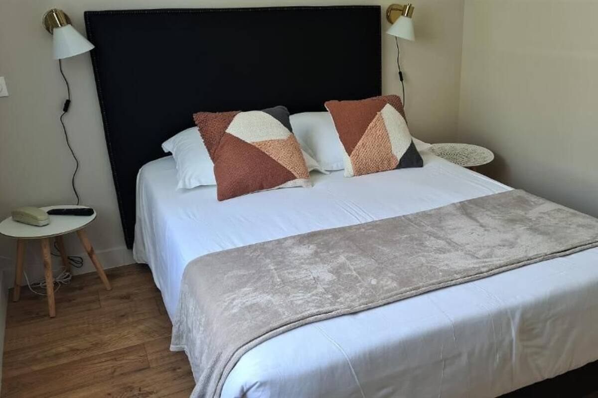 A neatly made bed with a white duvet, a grey throw blanket, and several patterned pillows. There is a bedside table with a lamp and a phone. The room has wooden flooring and a dark headboard.