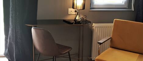 A room with a brown chair, a small table with a lamp, a window with curtains, and a radiator.