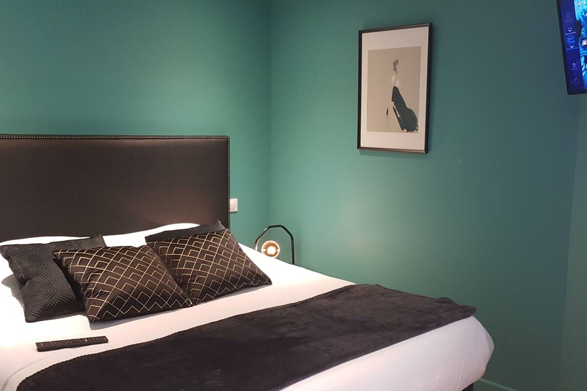 A bedroom with a bed, pillows, a bedside table with a lamp, a framed picture on the wall, and a TV displaying a blue screen.