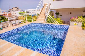 Piscina no terraço com azulejos de mosaico azul, cercada por um corrimão branco e uma escadaria para um andar superior.