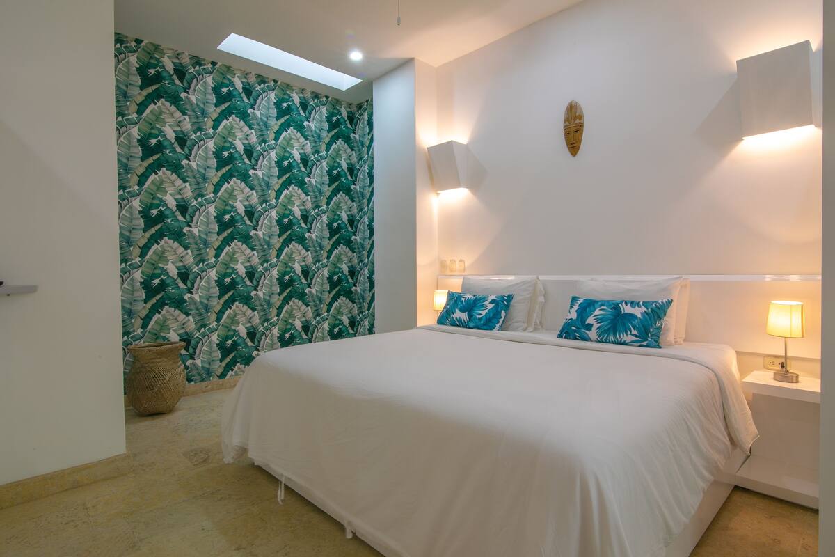 Une chambre avec un grand lit, une lucarne, un mur orné d’un motif végétal et un petit panier en osier.