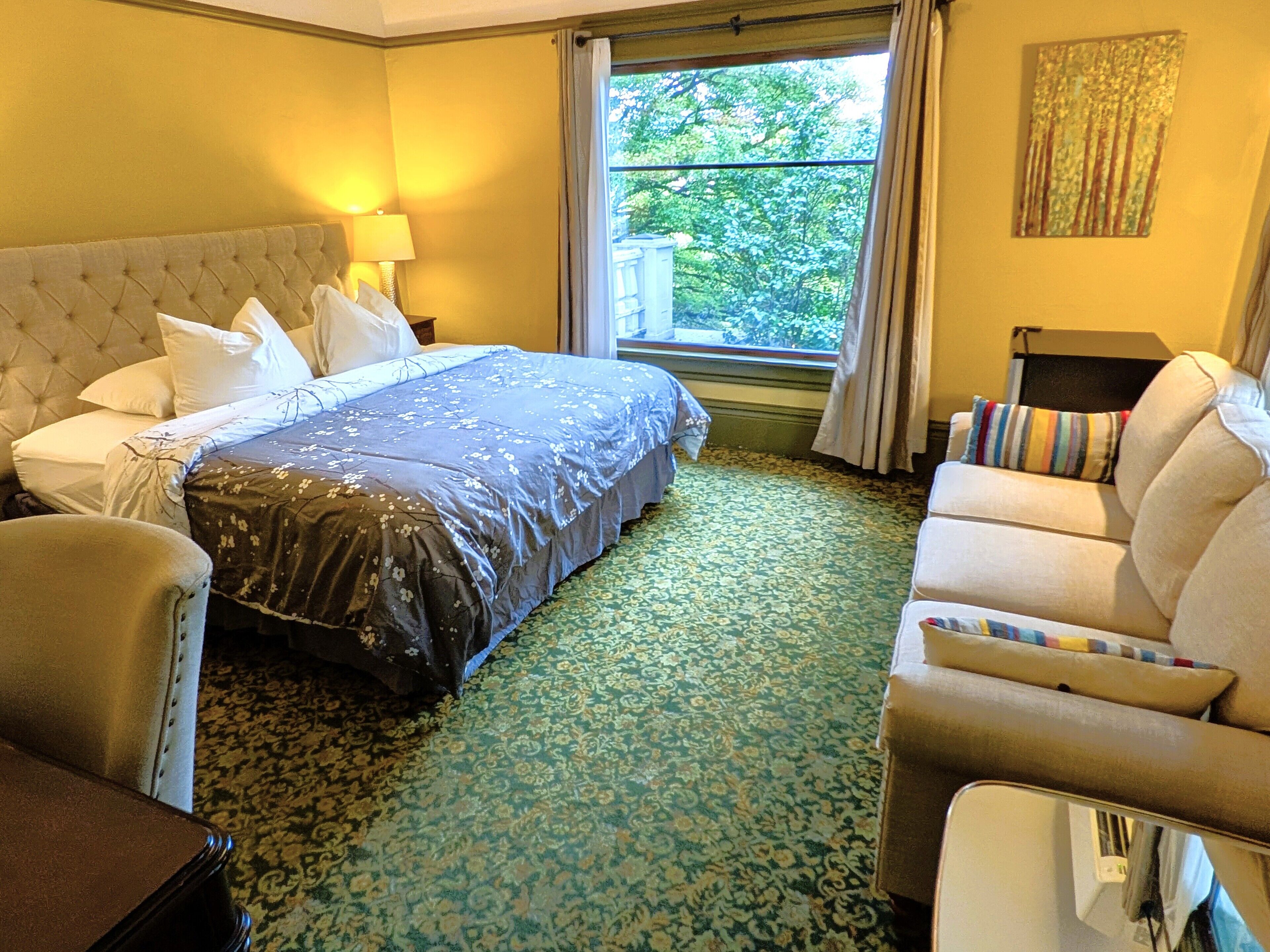 A hotel room with a large bed, a sofa, a chair, and a window with a view of trees.