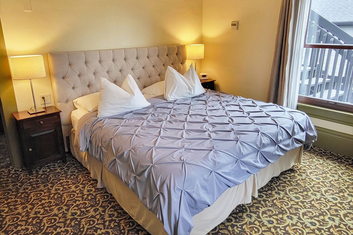 Une chambre d’hôtel avec un grand lit, deux lampes de chevet, une table de nuit, une moquette à motifs et une fenêtre donnant sur des immeubles.
