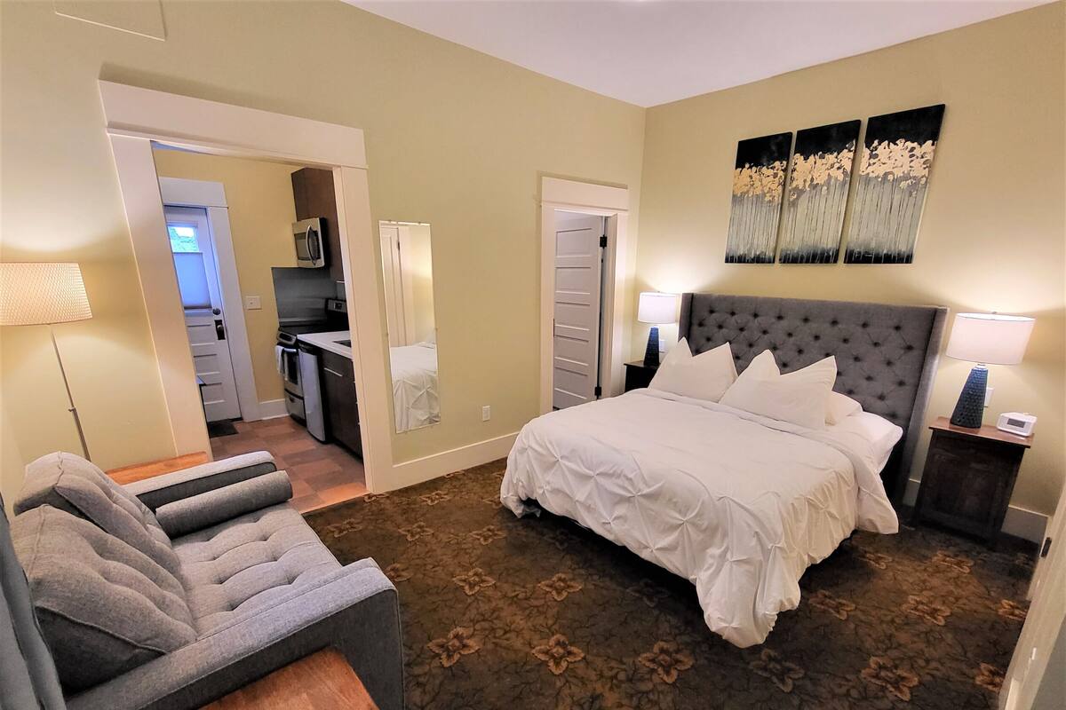 Une chambre d’hôtel avec un grand lit, un canapé gris, une petite table et un coin cuisine visible à travers une porte ouverte.