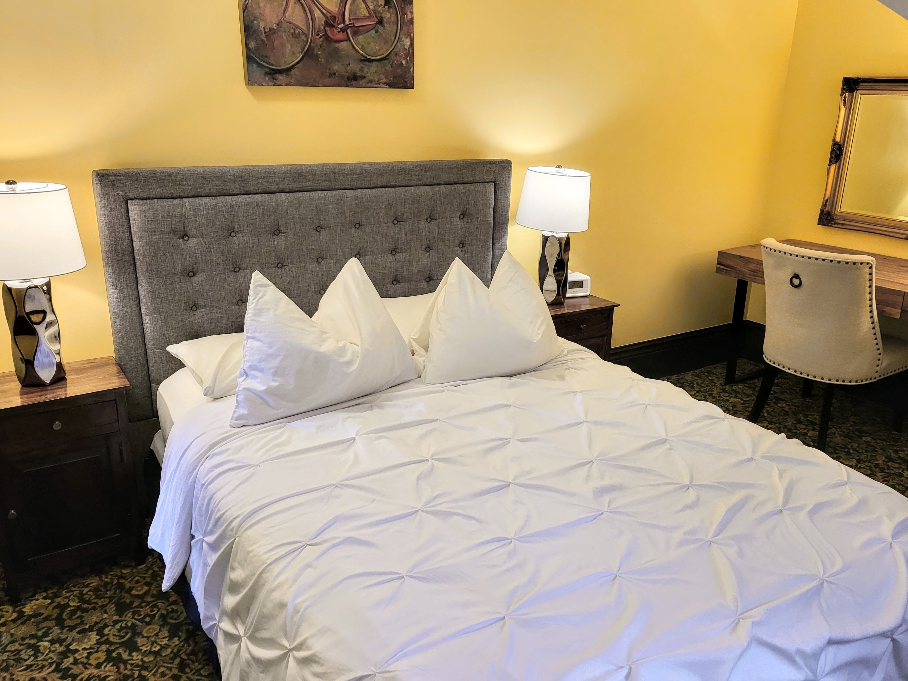 Un lit bien fait, doté d’une tête de lit capitonnée, deux tables de chevet avec des lampes, une chaise et un tableau accroché au mur.
