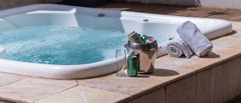 A hot tub with a tiled edge, a metal container holding a can and a glass, and a rolled towel on the side.
