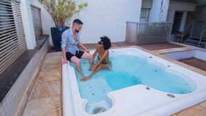 A hot tub with two people sitting inside, surrounded by a tiled patio area with a potted plant and a metal basket.