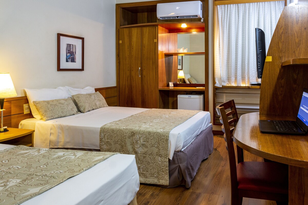 A hotel room with two beds, a desk with a laptop, a wardrobe, and an air conditioning unit.
