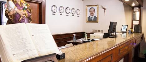 A reception desk with a wooden finish, a statue of a religious figure, multiple wall clocks, a framed portrait, and a bulletin board with notices.
