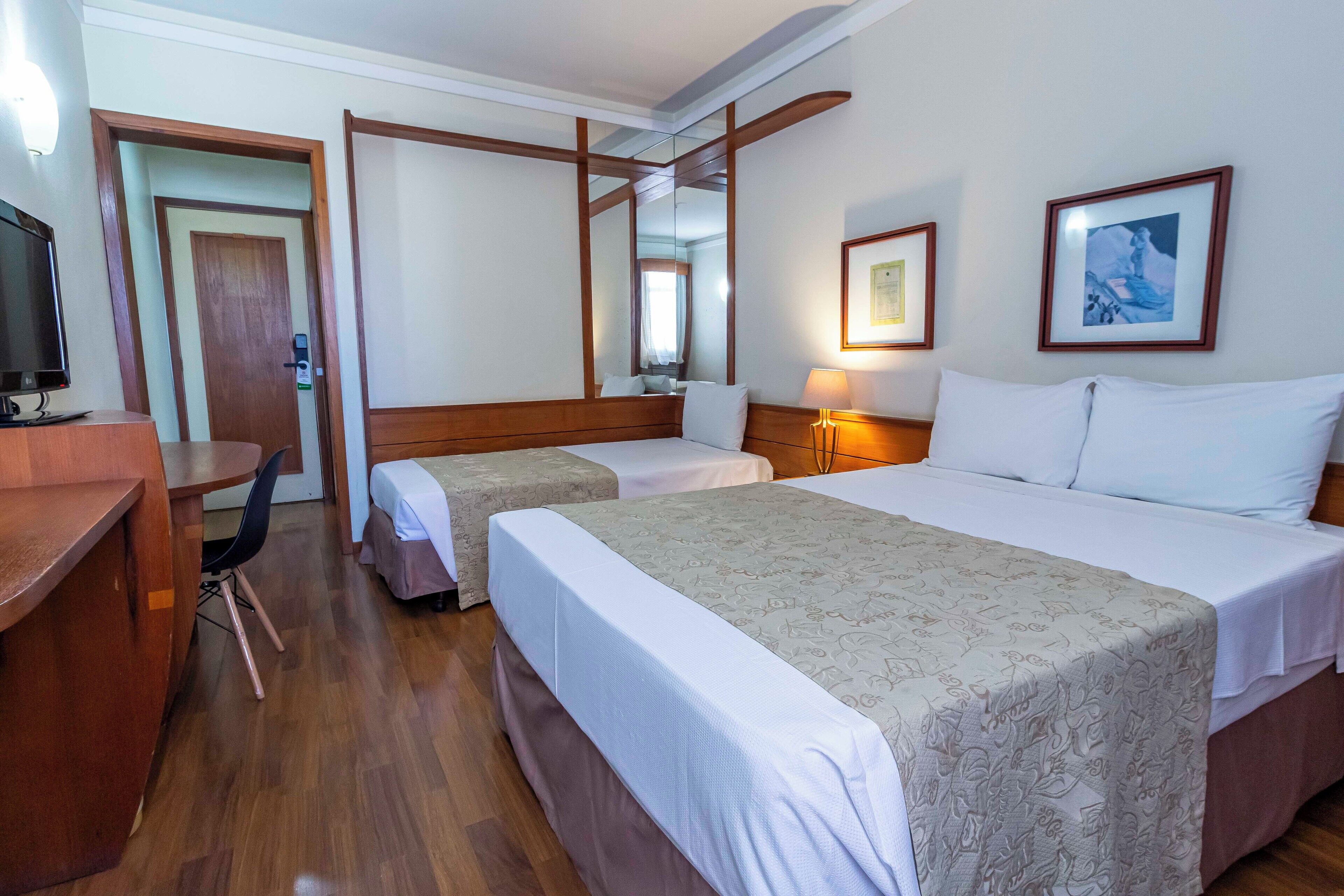 Quarto de hotel com duas camas, uma escrivaninha, uma cadeira, uma televisão e um espelho.