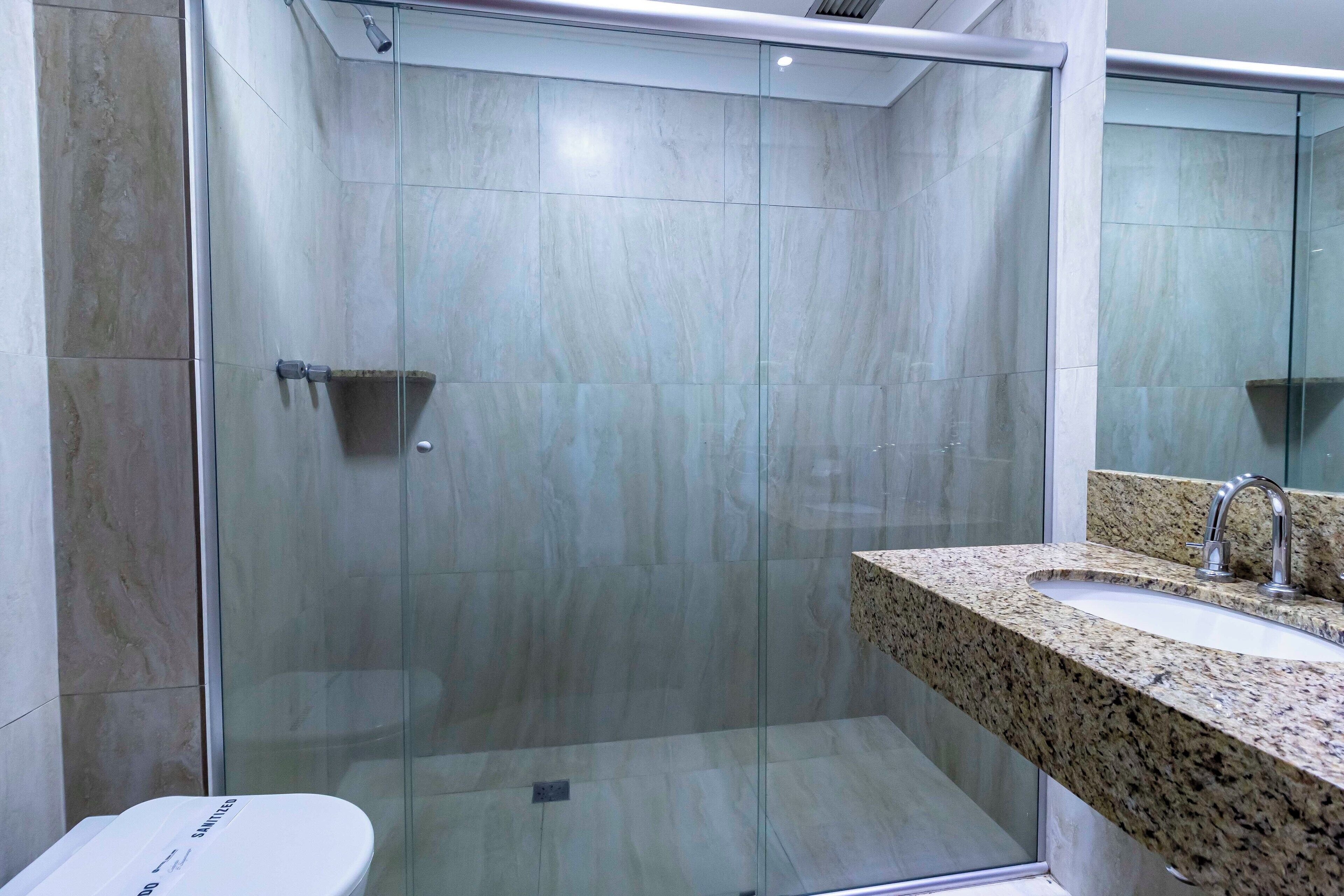 Un baño moderno con ducha envuelta en vidrio, lavamanos con encimera de granito y un inodoro con etiqueta visible.