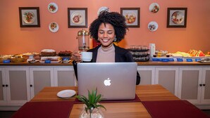 A person sitting at a table with a laptop, a coffee cup, and a small plant. The table is in a room with a buffet of food items and framed pictures on the wall. - Hotel Dan Inn São José do Rio Preto By Nacional Inn  (São José do Rio Preto)