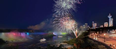 A nighttime cityscape with a prominent tower, a river with boats, and fireworks.