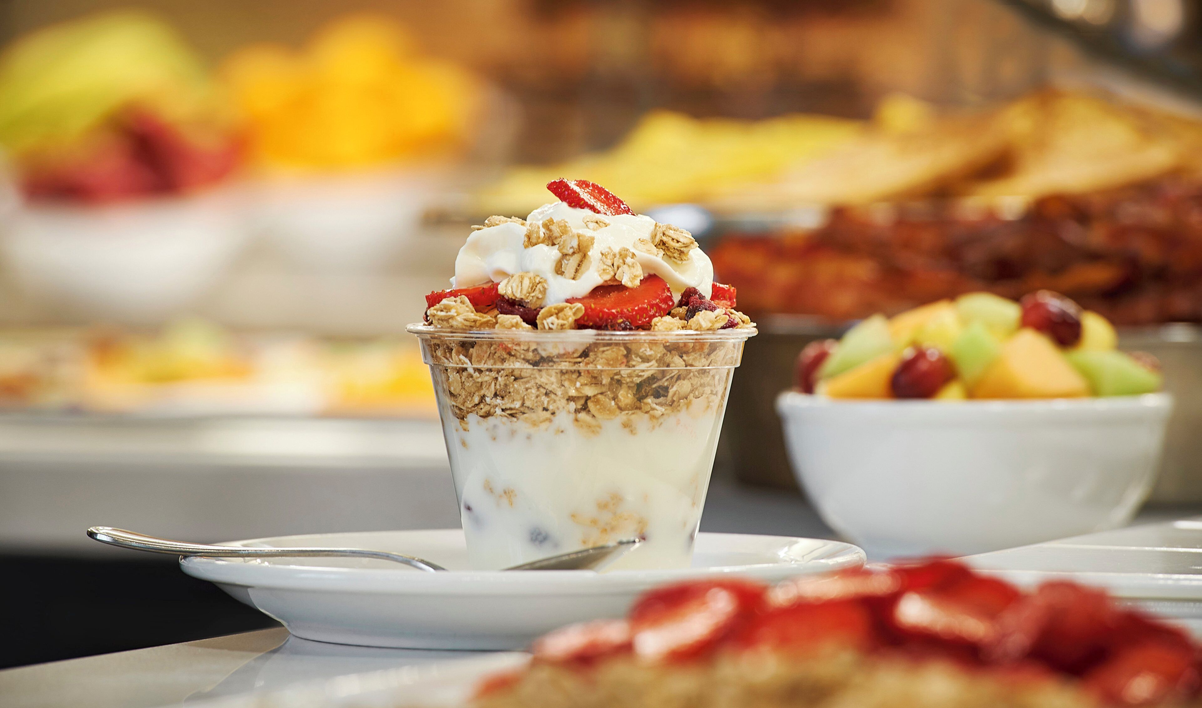 A dessert in a clear glass cup with granola, yogurt, and strawberries on a white plate.