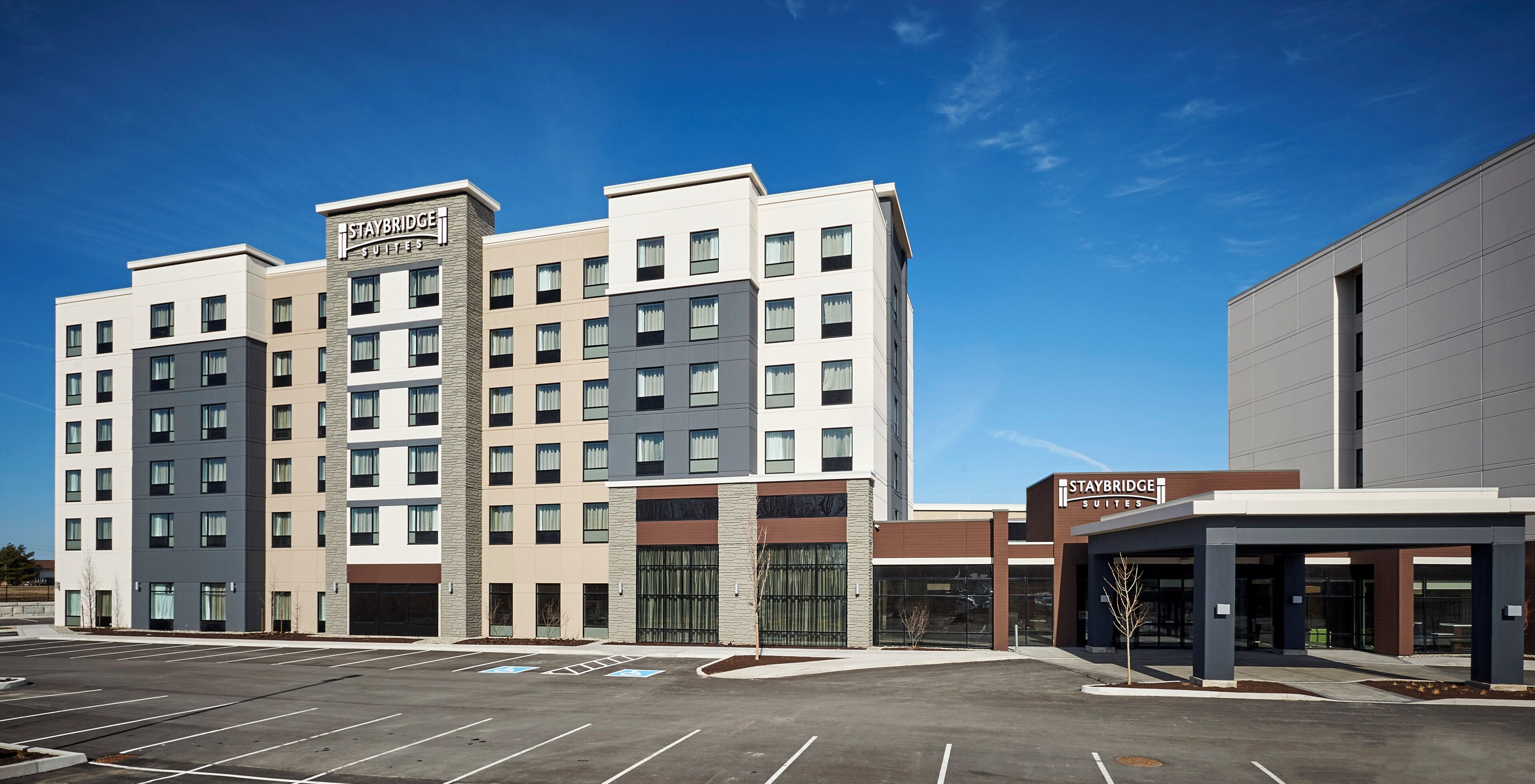 Un hotel de varios pisos con un letrero de Staybridge Suites, un estacionamiento y una fachada de edificio moderna.