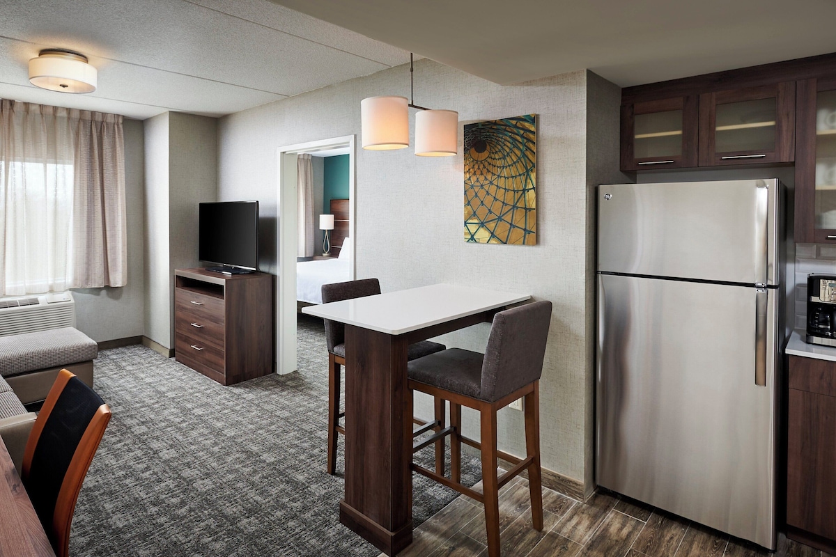 Een hotelkamer met een kitchenette, een zithoek met een tafel en stoelen, een bank, een televisie en een bed op de achtergrond.