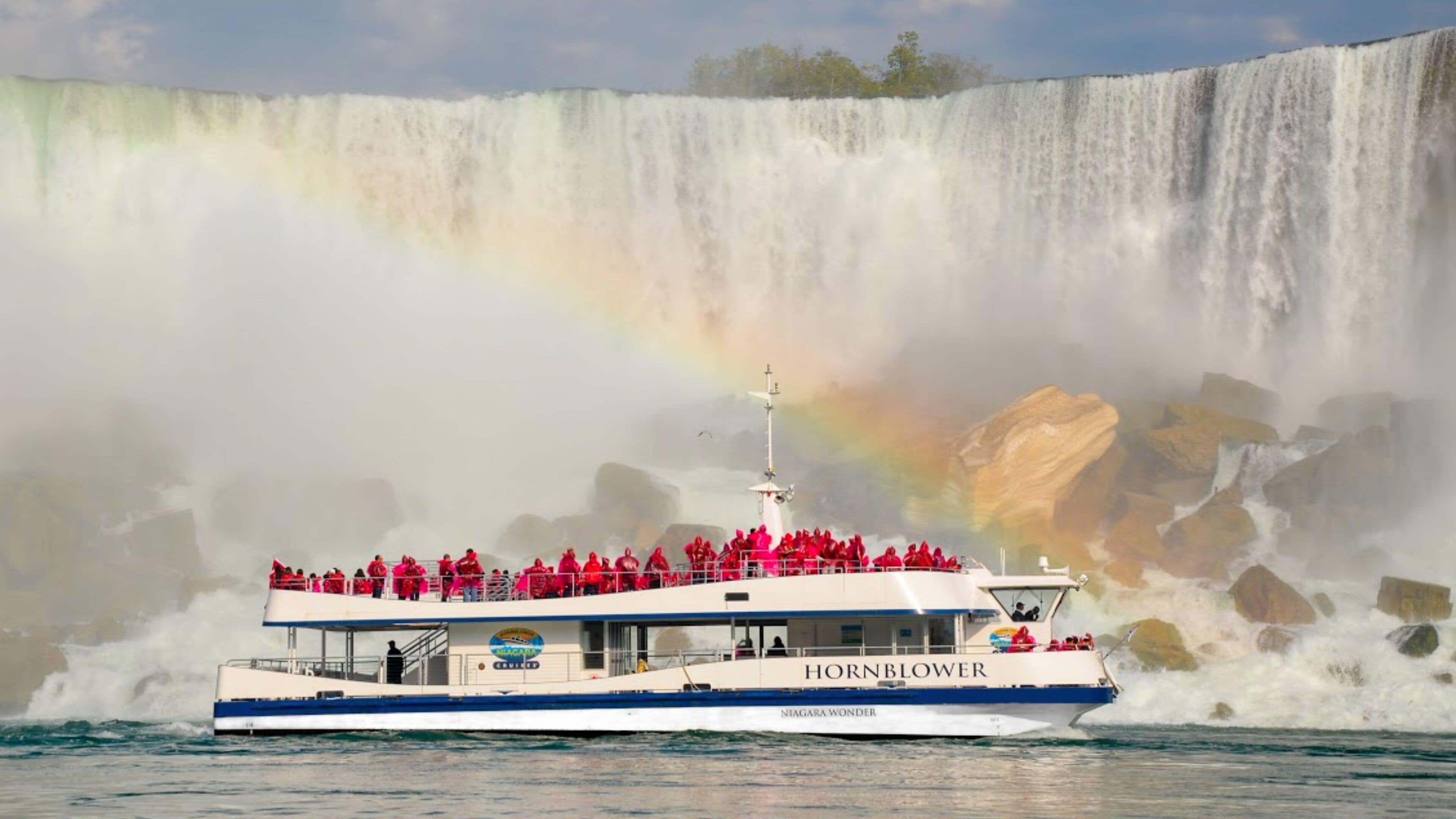 Ein Boot mit Passagieren in roten Jacken, beschriftet mit 'HORNBLOWER', vor einem Wasserfall mit einem Regenbogen.