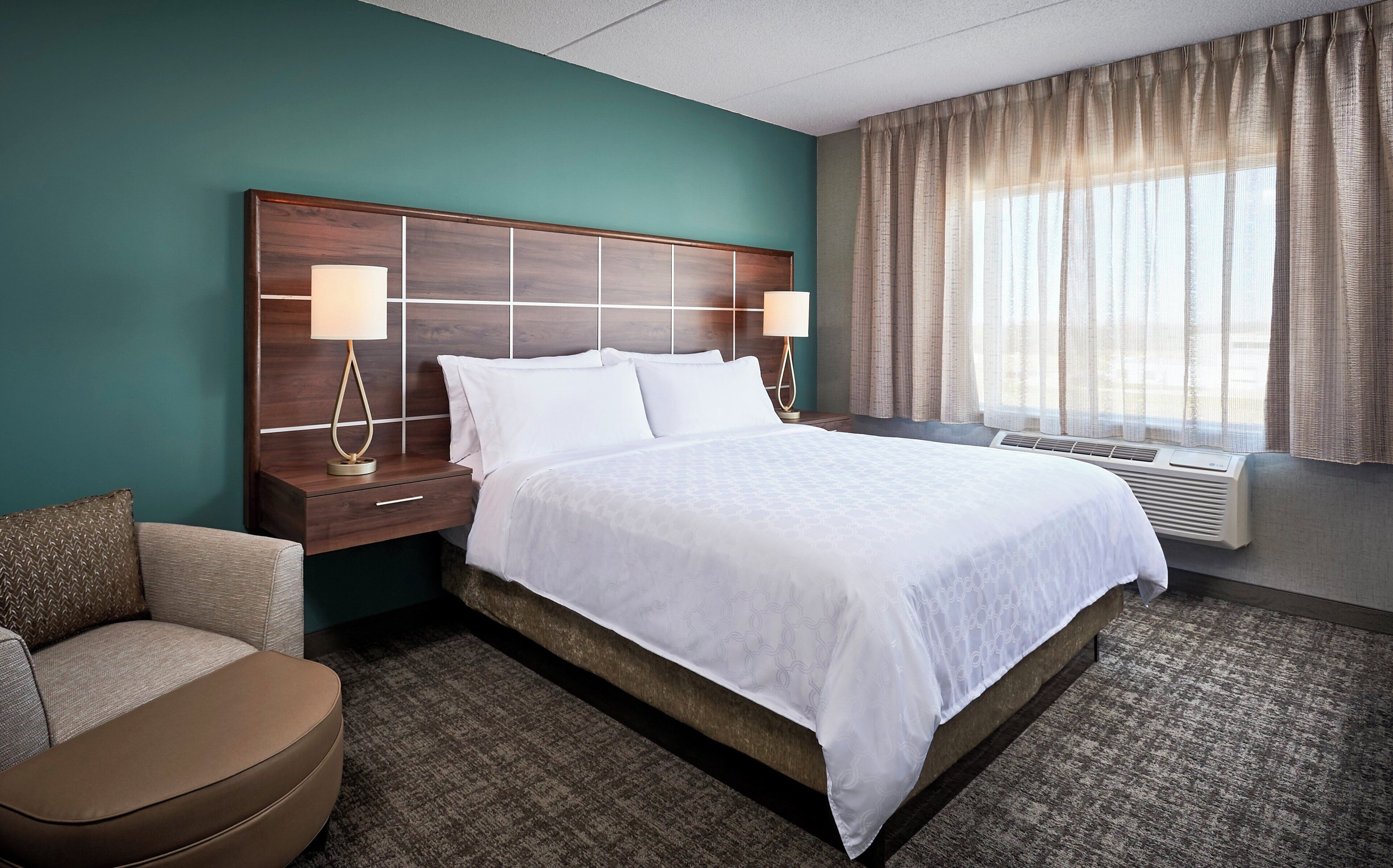 A hotel room with a large bed, two bedside lamps, a nightstand, a chair, and a window with curtains.