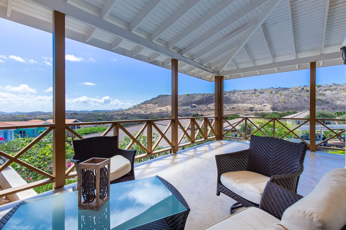 A covered outdoor seating area with wicker furniture and a glass table, overlooking a hilly landscape.