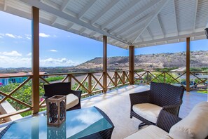 Un área de estar al aire libre con techo, mobiliario de mimbre y una mesa de vidrio, con vistas a un paisaje montañoso.