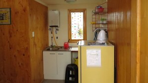 A compact kitchen with wooden walls, a window with colorful dots, a water heater, a kettle, and a refrigerator. - Cabanas Patricia en Horcon (Puchuncavi)