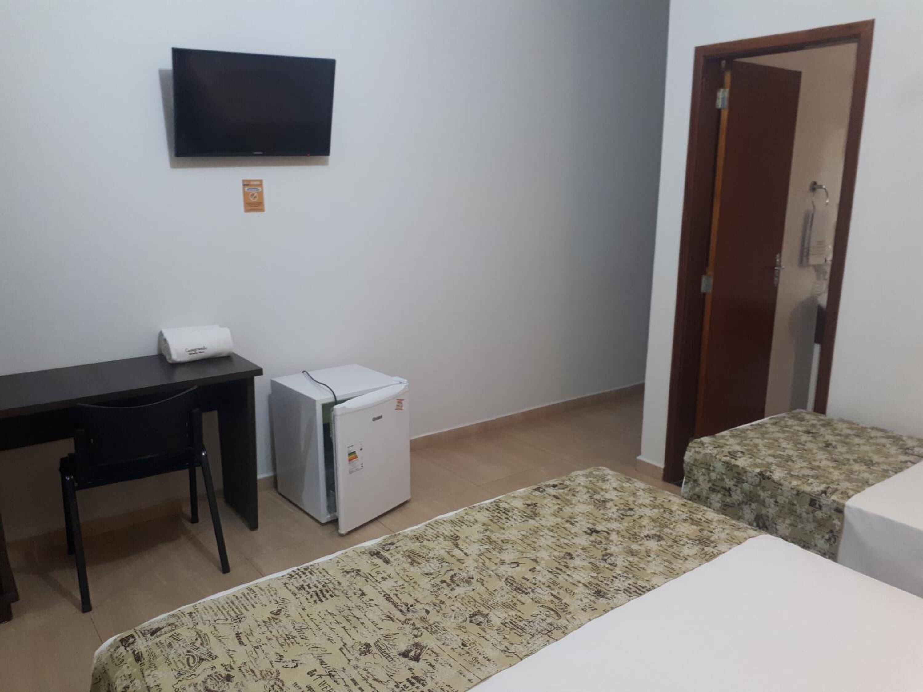 A room with a desk, a small refrigerator, two beds with patterned bedspreads, a television on the wall, and a door.