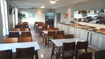 A cafeteria with multiple tables and chairs, a counter with food items, and a kitchen area in the background.