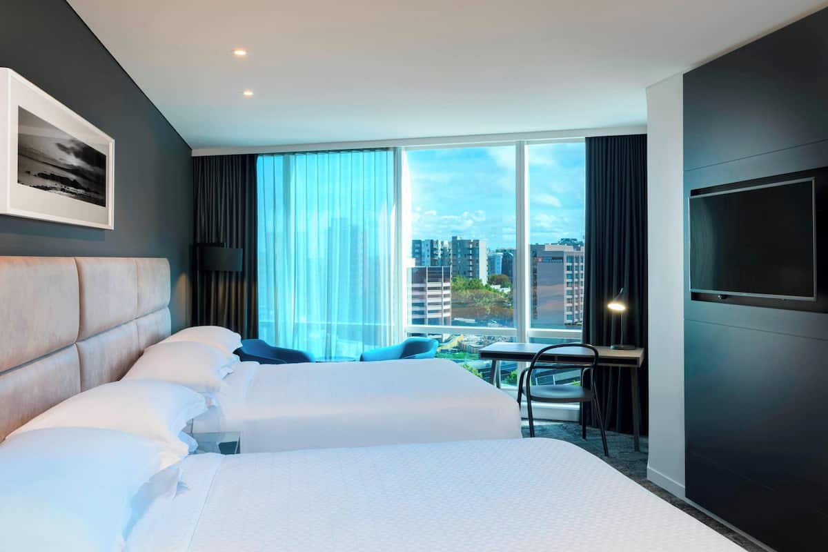 A hotel room with a large bed, a desk, and a TV. It has a city view through large windows.