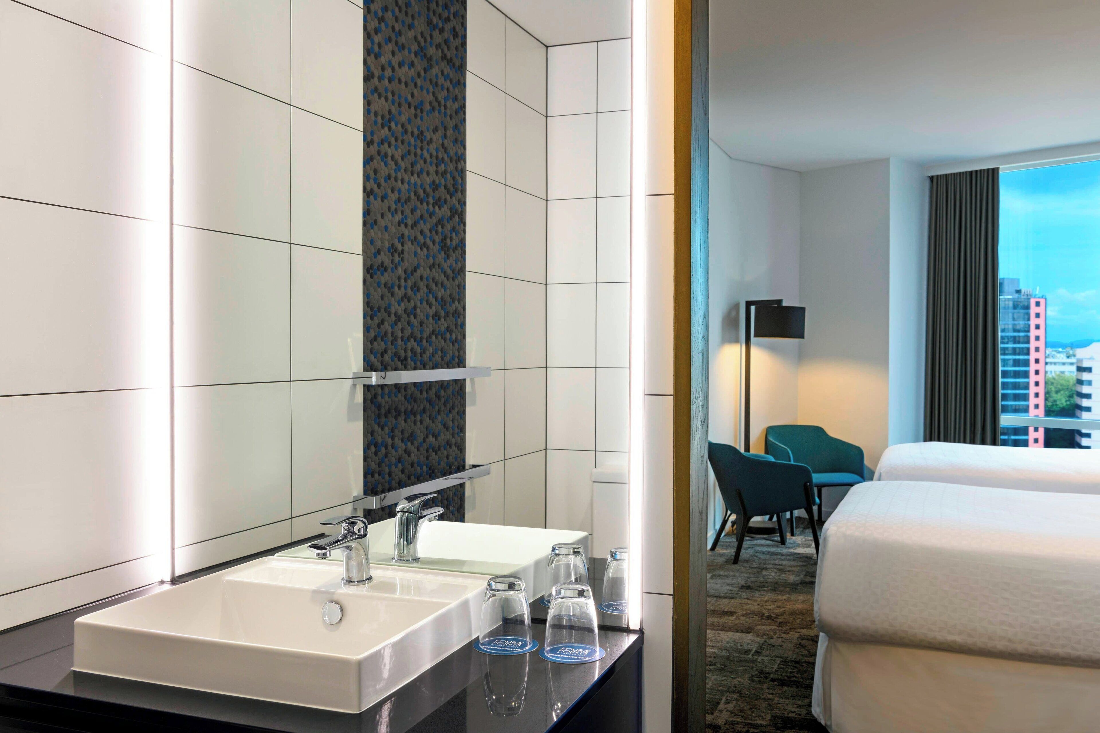 A hotel room with a large bed, a bathroom with a sink and mirror, and a view of the city through the window.