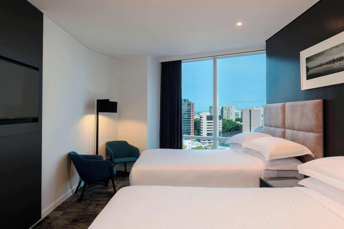 A hotel room with two beds, a desk, a chair, a large window with a city view, and a flat-screen TV.