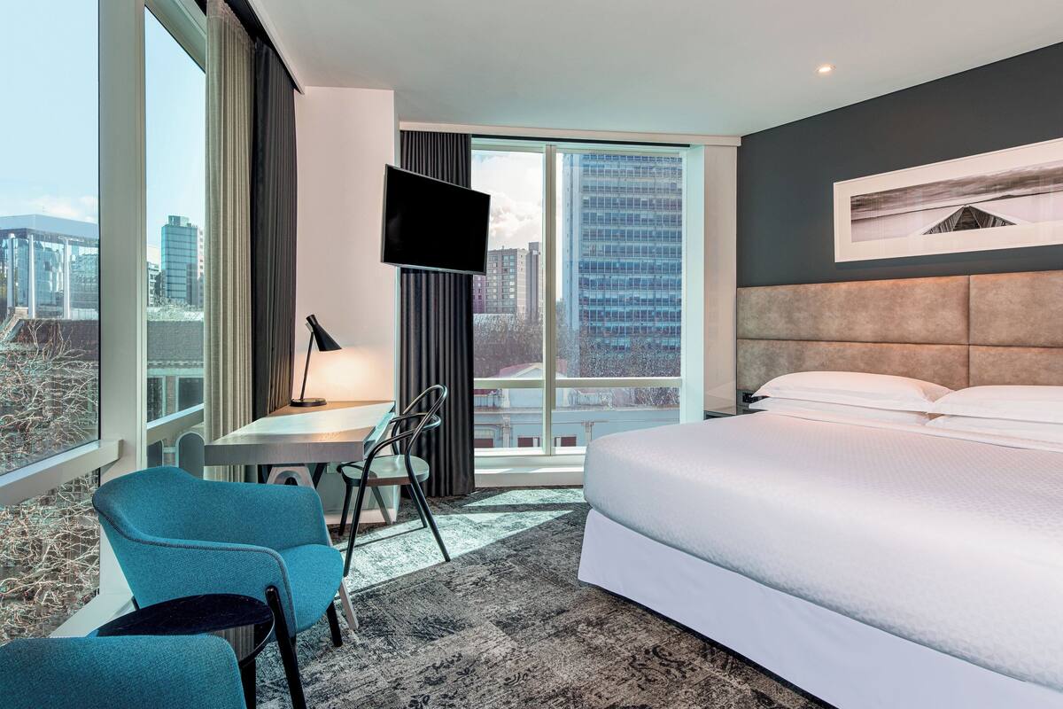 A modern hotel room with a large bed, a desk with a chair, a TV, and a city view.