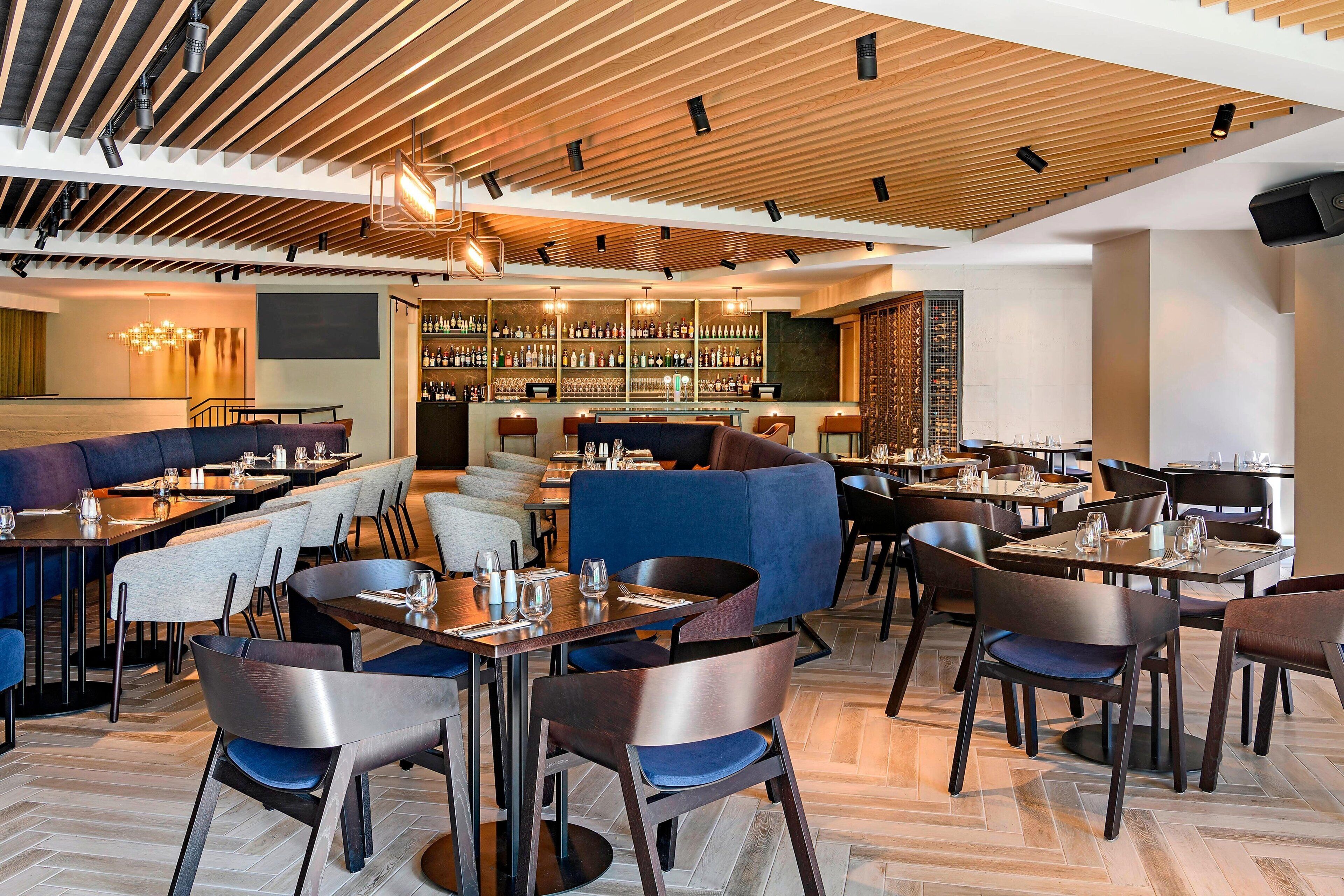 A modern restaurant with wooden ceiling beams, a bar area with shelves of bottles, and a mix of seating options including tables and booths.