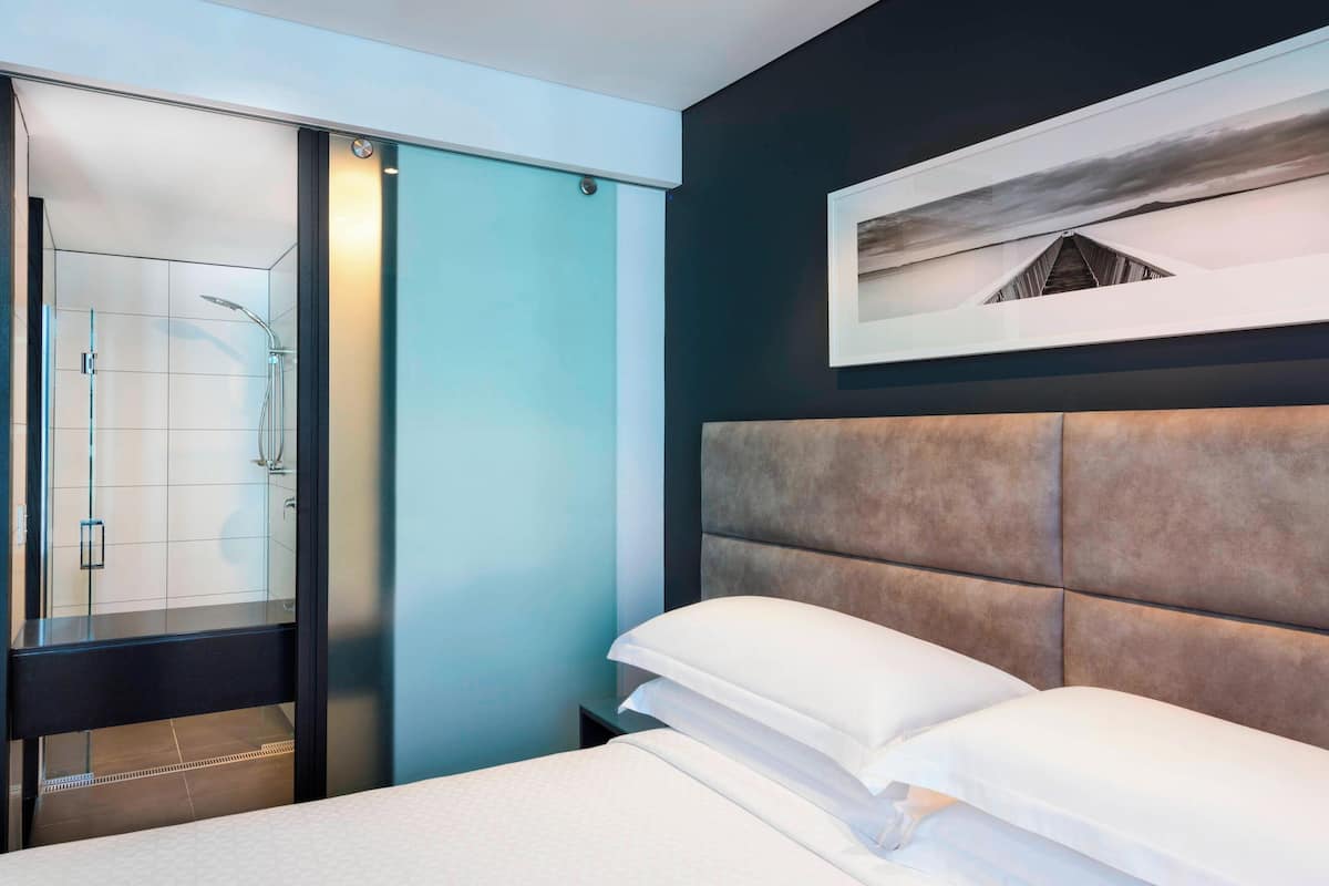 A modern hotel room with a glass shower enclosure, a large bed with white linens, and a framed artwork on the wall.