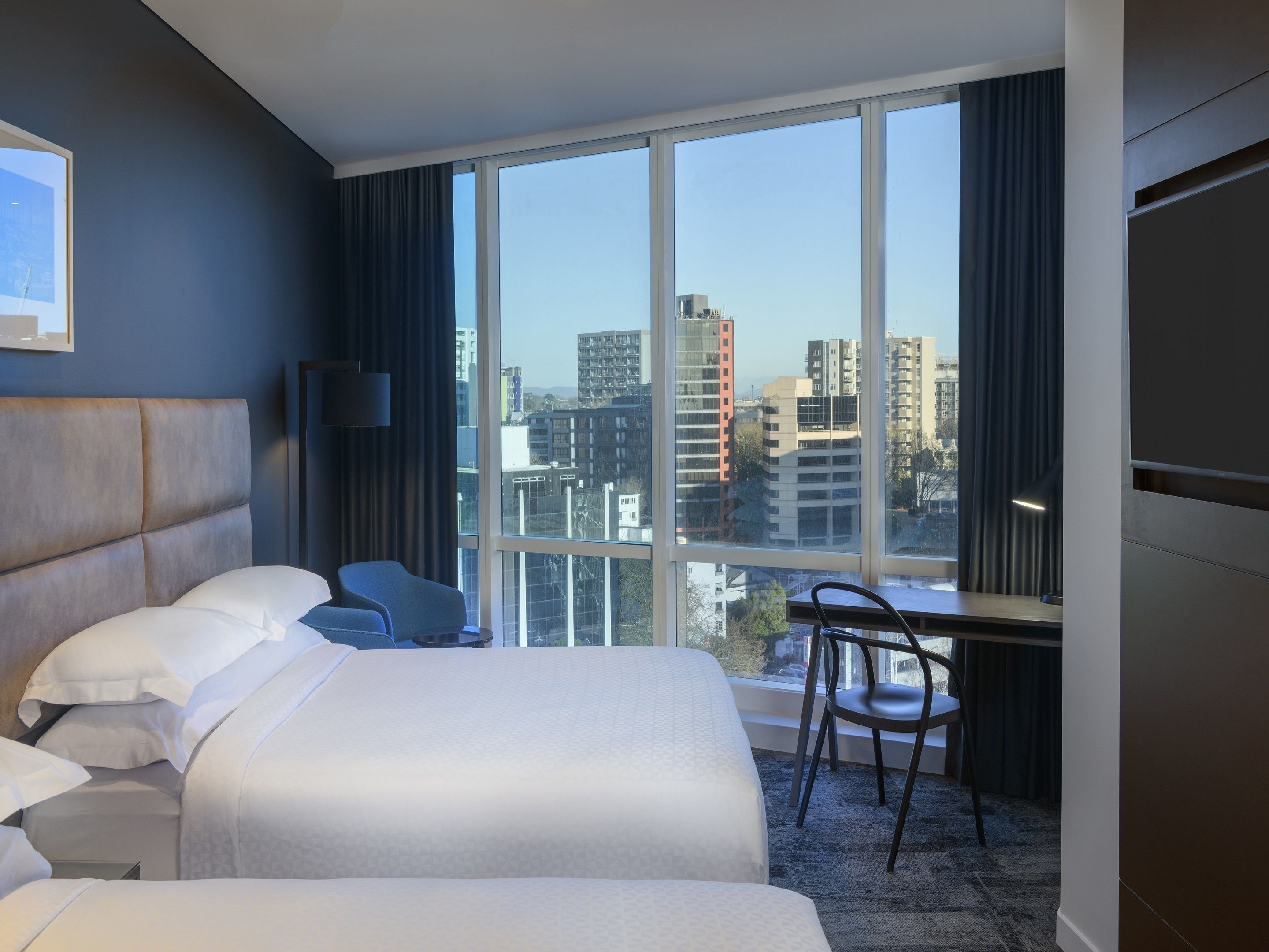 A hotel room with a large bed, a desk, a chair, and a view of the cityscape through large windows.