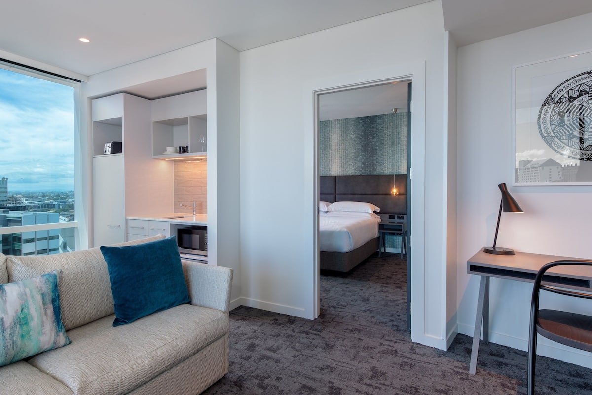 A modern hotel room with a sofa, a desk, a bed, and a view of the city.